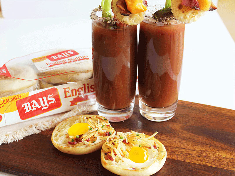 BAYS WITH EGGS AND BACON NEXT TO A RED DRINK WITH CELERY CHEESE AND BACON AS A GARNISH
